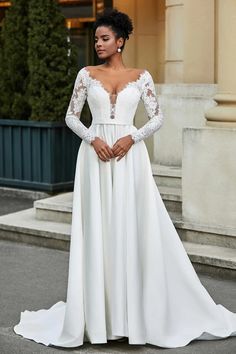 a woman wearing a white wedding gown with long sleeves and lace on the top, standing in front of a building