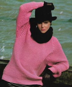 a woman in a pink sweater and black hat leaning on a rock by the water