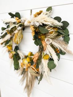 a dried wreath hanging on the side of a white wall with green leaves and yellow flowers