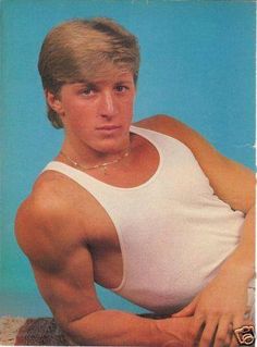 the man is posing with his arms crossed and wearing a white tank top, blue background