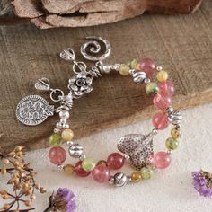 This artisan handmade double-strand bracelet showcases the natural beauty of beautiful grade gemstones, including Strawberry Quartz (10.8mm), Rutilated Quartz (6.8 mm), and Prehnite (8 mm), that carefully selected to make the colors of this bracelet stand out, creating a harmonious blend of pink, green, and yellow hues. The bracelet is adorned with intricate Karen Hill Tribe fine silver accents, featuring charming elements like a hammered heart-shaped bead, flower S-clasp, and unique charms, Including 925 sterling silver, all expertly strung together with a strong and durable waxed cord, finished with woven ends and Hill Tribe fine silver chain. Whether you're looking to add a statement piece to your jewelry collection, attend a festival or a boho-themed event, search for a thoughtful gift Agate Bracelet With 108 Beads, Fusion Style Beaded Bracelets With Gemstones, Bohemian Tourmaline Jewelry With Natural Stones, Artisan Bracelets With Natural Stone Round Beads, Artisan Bracelets With Round Natural Stone Beads, Artisan Gemstone Beads Round Bracelets, Handmade Fusion Beaded Bracelets For Healing, Bohemian Double Strand Beaded Bracelets For Jewelry Making, Artisan Bracelets With Natural Stones