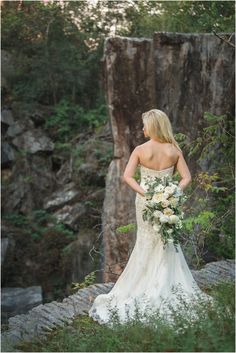 a woman in a wedding dress is standing on rocks and looking down at her bouquet