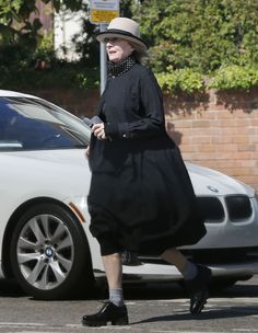 an older woman wearing a black dress and hat crosses the street in front of a white car