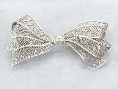 Add a touch of elegance and whimsy to any outfit with this bow brooch. The delicate filigree detailing and sparkling European Cut diamond make this brooch a timeless statement piece. Perfect for any occasion!Metal: 14K White GoldGem: European Cut Diamond .10 Carats, SI1 in Clarity, I in ColorGem Measurements: 3.2 mm, RoundMeasurements: 44 x 23 mmMarks: "X14K" Stamped on the pin guard Classic Wedding Brooches With Intricate Design, Elegant White Gold Brooch With Intricate Design, White Gold Brooches With Intricate Design For Wedding, Elegant White Gold Brooches With Intricate Design, Wedding Brooch With Intricate Diamond Design, White Gold Wedding Brooches With Intricate Design, Elegant Brooches With Intricate Design For Anniversary, Elegant Filigree Brooches For Evening, Wedding Diamond Brooch With Intricate Design