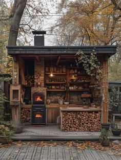 an outdoor kitchen with wood stacked on the outside and fire in the fireplace, surrounded by trees