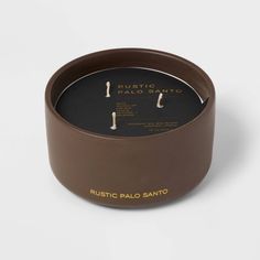 a candle that is sitting inside of a brown container on a white surface with the words rustic palo sano written in gold