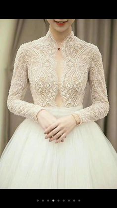 a woman wearing a white dress with long sleeves and beading on the neck, standing in front of a curtain