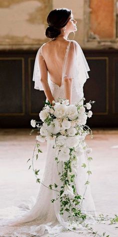 a woman in a wedding dress holding a bouquet of white flowers and greenery with her back to the camera