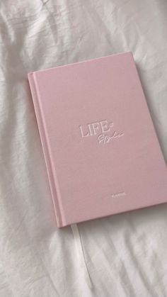 a pink book sitting on top of a white sheet covered bed next to a pillow