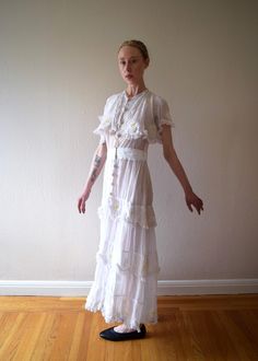 An antique Edwardian era from the 1910s. It is a really unique dress with embroidered daisies. It also has a ruffled overlayer on the bodice, a skirt with tiered ruffles, inset lace, and lace trim. The dress has decorative crochet-covered buttons and fastens with snaps and hook and eyes up the front. The dress has two kinds of fabric -- a lightweight cotton as well as a cotton net. Size: xs to small Bust: up to 34" Waist: 26" Hip: 42" (meant to drape) Length: 54-55" Condition: Very good. Faint yellowing on front waistband that didn't completely lift when I cleaned dress. A handful of small hand repairs -- the two pictured are on the netting above back waist and on the back shoulder. None of the flaws jump out at you, and the dress presents beautifully. Fitted Tiered Dress With Lace Trim, Tiered Lace Maxi Dress With Fitted Bodice, White Fitted Victorian Dress For Summer, White Fitted Victorian Summer Dress, Fitted White Vintage Dress With Ruffles, White Fitted Vintage Dress With Ruffles, White Fitted Ruffled Vintage Dress, White Victorian Ruffled Dress, White Victorian Dresses With Ruffles