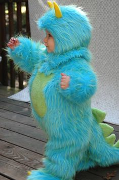 a small child dressed in a blue monster costume sitting on a wooden deck with his arms outstretched