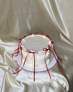 a white cake with red ribbon around it sitting on a satin tablecloth covered surface