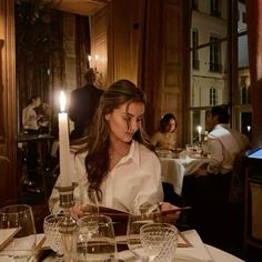 a woman sitting at a table with wine glasses and candles in front of her, reading a book