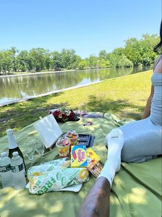 a woman sitting on a blanket next to a lake with food and drinks in front of her