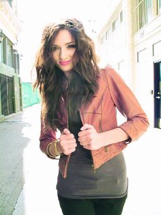 a woman is standing on the street with her hands in her pockets and wearing a brown leather jacket