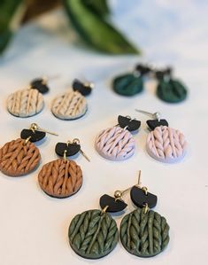 six pairs of earrings are shown on a white surface with green leaves in the background