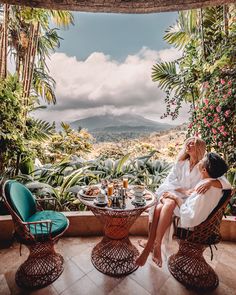 two people sitting at a table in front of some plants and trees with mountains in the background