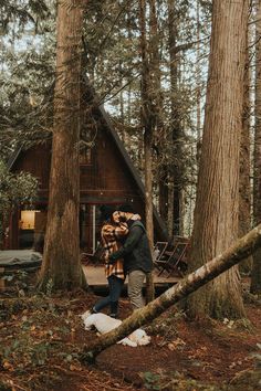 a man and woman standing in front of a cabin surrounded by tall trees with their arms around each other