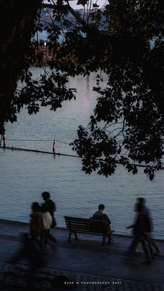 people are sitting on a bench near the water at sunset or dusk, with one person walking in the distance