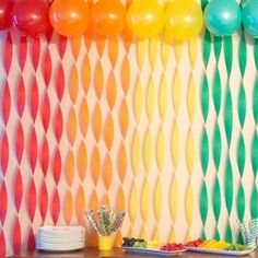 a rainbow party with balloons and streamers on the wall, plates and cups in front of it