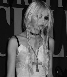 a woman with long blonde hair wearing a cross necklace and black eyeliners is standing in front of a wall