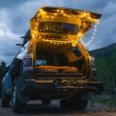 A parked SUV with its tailgate open, revealing a cozy setup with a mattress inside. Luno® Camping String Lights are draped around the tailgate, emitting a soft glow with adjustable brightness. The vehicle is surrounded by trees under a cloudy, dusk sky, powered by a rechargeable USB-C battery pack. Barn Door Window Covering, Camping String Lights, Firewood Carrier, Barn Door Window, Door Window Covering, Camping Fan, Slider Door, Shoe Storage Bags, Camping Pillows