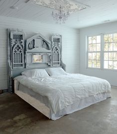 a white bed sitting under a chandelier in a bedroom next to a window