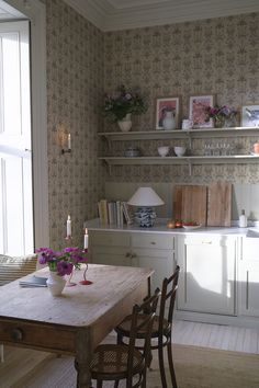 a dining room table and chairs in front of a wallpapered kitchen with open shelving