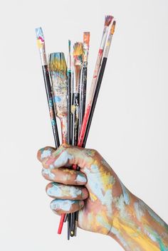 a person's hand holding paintbrushes with multiple colors on their body and hands