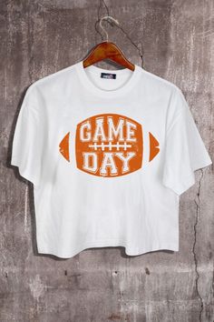 This White Orange Game Day Football Graphic Crop Tee is tailored for comfort and style. It is made from soft, lightweight material with a loose fit and crew neck design. Its unique football graphic adds a fun touch to any casual look. Fabric & fit: 100% cotton Model is wearing size Small. Game Day Football Season T-shirt With Logo, Sporty T-shirt With Football Season Screen Print, Sporty T-shirt For Football Season Game Day, Sporty T-shirt For Game Day Football Season, Sporty T-shirt For Game Day During Football Season, White Sporty T-shirt For Football Season, White Screen Print T-shirt For Football Season, White T-shirt With Football Season Screen Print, Relaxed Fit Letter Print T-shirt For Football Season