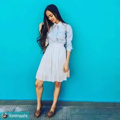 a woman standing in front of a blue wall wearing a white dress and brown shoes