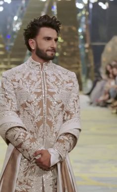 a man in a white and gold outfit standing on a runway with other people watching