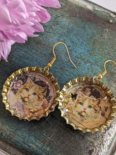 a pair of earrings with pictures of cats on them sitting next to a pink flower