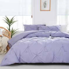 a bed with purple comforter and pillows in a room next to a plant on the floor