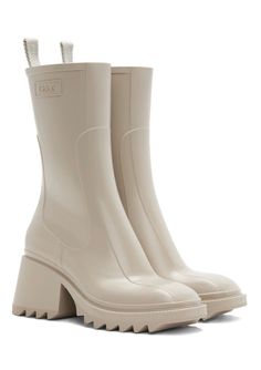 The rubber Betty rain boot reveals a sleek, strong upper recalling classic leather boots and incorporating the Maison's signature contours. The voluntarily oversized back loop shows the Chloé logo, while an internal zipper closure ensures a fitted, feminine silhouette. Short and flat, the square toe shape lends a strong, assertive attitude. The comfortable heel elevates the silhouette, while the bold lugged sole gives an edgy attitude to this rain boot. Ankle Boots For Rainy Weather In Fall, Ankle Boots For Rainy Weather And Fall, Rainy Weather Ankle Boots, Chloe Rain Boots, Chloe Betty Boots, Pvc Boots, Chloe Logo, Chloe Clothing, Beige Boots