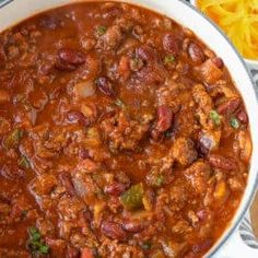 a large pot filled with chili and meat on top of a wooden table next to cheese wedges