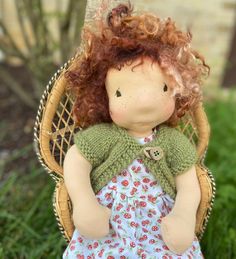 a doll with red hair sitting in a wicker chair on the grass next to a tree