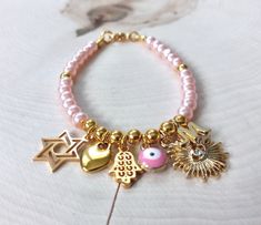 Handcrafted wire-strung beaded charm bracelet designed with 4mm pearlized glass in baby girl pink with your choice of gold plated charms with lobster claw clasp. A lovely keepsake for Baby Naming, new birth, Zeved Habat, First Siddur; or, in larger sizes for Bat Mitzvah; for adults for expectant mother, Baby Shower, Wedding, Bride, Mother of the Bride, Bridesmaids Favors, New Mom, Mother/Child or any religious faith and family celebration. Bracelet is also available in white, baby blue or taupe. CHOOSE YOUR GOLD CHARMS: All-Seeing Eye (5/8" gold plated micro pave) Birthstone (3/8" stainless gold plated: PLEASE INDICATE BIRTH MONTH) Blue Evil Eye (1/4" gold plated with enamel) Crescent Moon (3/8" gold plated micro pave) Evil Eye (3/8" gold plated micro pave) Hand Of God Hamsa (3/8" gold pla Spiritual Pink Jewelry For Birthday, Pink Metal Beaded Bracelet With Charms, Pink Metal Beaded Bracelets With Charms, Pink Hypoallergenic Charm Bracelet Gift, Handmade Pink Gold Beaded Bracelets As Gift, Handmade Pink Gold Beaded Bracelet As Gift, Spiritual Gold Beaded Bracelets With Charms, Pink Charm Bracelet For Birthday, Adjustable Pink Nickel-free Charm Bracelet