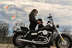 a man and woman standing next to a motorcycle on a dirt road with clouds in the background