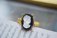 This ring features an elegant gold band with a classic cameo design. The centerpiece is an oval-shaped stone with a deep black background, showcasing the profile of a woman's face in white relief. The setting around the stone has intricate gold detailing, adding to its vintage charm. Details Isabella Black Cameo Vintage Ring Adjustable opening Gold color Rare, antique design Reyis Jewelry Isabella Collection Black Cameo, Pearl Jewelry Necklace, Natural Stones Necklace, Gold Statement Earrings, Natural Stone Jewelry, Natural Stone Bracelets, Antique Design, Deep Black, Anklet Jewelry