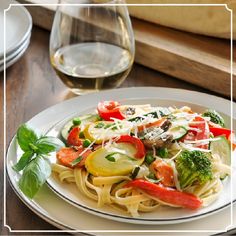 a white plate topped with pasta and veggies next to a glass of wine