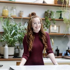 a woman standing in front of a counter with plants on the shelves behind her and smiling at the camera