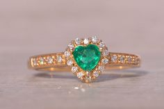 The Mademoiselle: Heart Shaped Natural Emerald and Diamond Ring in Yellow Gold. This exquisite ring features a heart brilliant-cut natural emerald, measuring 4.8mm by 4.7mm, showcasing a vivid very slightly bluish-green color. The emerald is surrounded by a halo of round brilliant-cut natural diamonds, which extend down the shank, enhancing the ring's brilliance. Expertly crafted in 14-karat yellow gold, this piece is currently a size 7.25 but can be resized to fit any finger for an additional f Elegant Heart-shaped Green Emerald Ring, Elegant Heart-shaped Emerald Ring For Anniversary, Elegant Green Heart-shaped Emerald Ring, Elegant Emerald Heart-shaped Ring, Elegant Heart Shaped Emerald Ring For May Birthstone, Elegant Heart-shaped Emerald Ring For May Birthstone, Elegant Heart Cut Green Emerald Ring, Elegant Green Emerald Heart Cut Ring, Elegant Green Heart Cut Emerald Ring