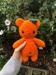 a hand holding an orange crocheted teddy bear in front of some bushes and flowers