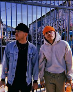 two young men standing next to each other in front of a fence with graffiti on it