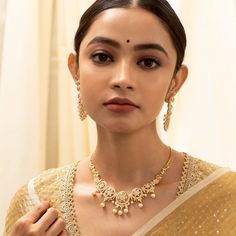 a woman wearing a gold necklace and matching earrings with her hand on her shoulder, looking at the camera