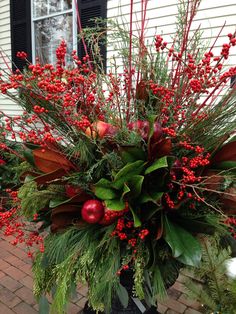 a vase filled with red berries and greenery on top of a brick floor next to a house