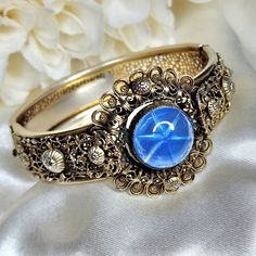 Stunning Art Deco filigree hinged bracelet circa 1930's. Beautiful gold tone lacy filigree metal work and a large blue glass Cabochon. Clasp fastens snugly. Inside measurement is approximately 7¼ inches. There is some wear to the gold plating consistent with age. Please refer to the photos. In overall wonderful vintage condition. Luxury Ornate Cabochon Bracelets, Art Deco Cabochon Jewelry For Anniversary, Formal Brass Cabochon Jewelry, Formal Brass Jewelry With Cabochon, Gold Cabochon Bangle Jewelry, Antique Gold Cabochon Bracelets, Vintage Metal Jewelry With Intricate Design, Antique Gold Bracelets With Cabochon, Engraved Heirloom Metal Jewelry