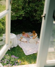 two women and a baby are sitting on a blanket in the grass near an open window