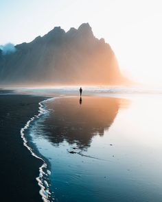 a lone person standing on the beach in front of mountains and water at sunset or sunrise